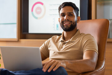 Smiling man using laptop in office with charts on screen in background