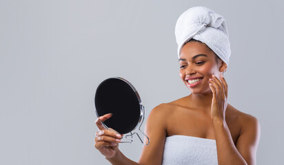 Satisfied smiling black woman wrapped in towel checking her face in mirror after shower over grey background, skincare concept