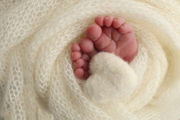 Obraz premium The tiny foot of a newborn baby. Soft feet of a new born in a white wool blanket. Close up of toes, heels and feet of a newborn. Knitted white heart in the legs of a baby. Macro photography. 