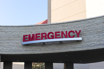 Obraz premium Red EMERGENCY sign marks the entrance to a hospital emergency department, mounted on a curved stone façade beneath a canopy structure, signaling critical care access for arriving patients
