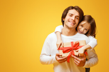 Cute little daughter and father holding present, girl hugging dad from behind, Father Day concept, yellow background