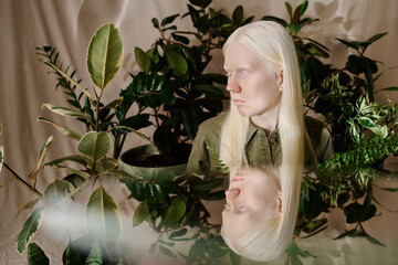 Young woman with albinism and her mirror image in studio decorated with green houseplants, conceptual portrait