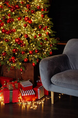 A Christmas tree with red and green decorations and presents under it stands in front of a gray sofa. No people.