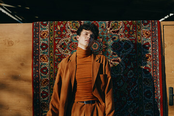 Stylish young man in a rust colored outfit poses against a vibrant patterned rug backdrop