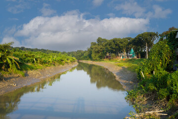 Fototapeta premium Blue sky above, clear narrow river below. Temple by the river. Image of green Bangladesh in Asia.