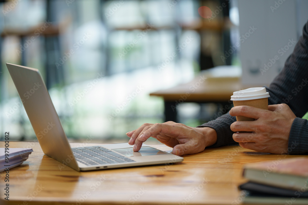 Poster a man is typing on a laptop computer while holding a cup of coffee. the scene suggests a busy work e