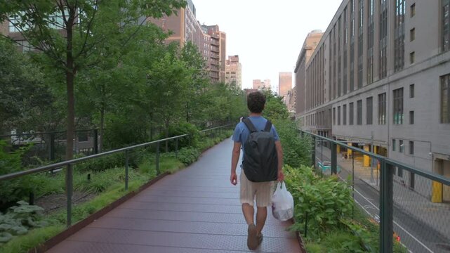 New York City, USA, August 14, 2024. POV footage walking along the high line: a young Caucasian man precedes us, he is walking among the green trees. Below we can see cars passing by.