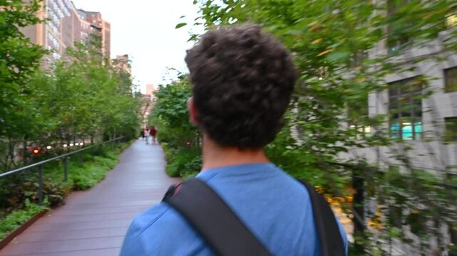 New York City, USA, August 14, 2024. POV footage walking along the high line: a young Caucasian man precedes us, he is walking among the green trees. Below we can see cars passing by.