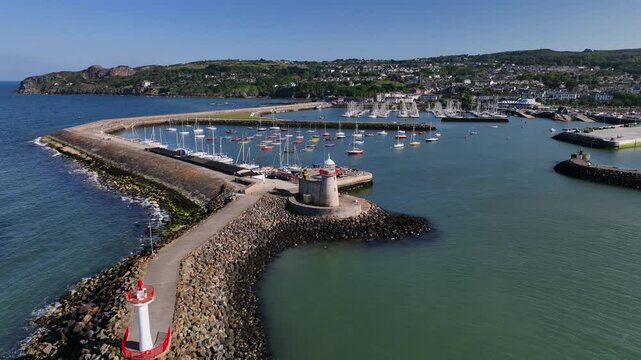 Howth Harbour, County Dublin, Ireland, May 2023. Drone orbits counter clockwise around the Lighthouse at the mouth of the harbour on a bright sunny afternoon with Ireland's Eye in the background.