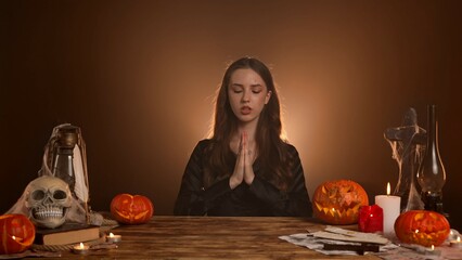 Halloween gothic woman witch fortune teller sitting at table holding hands in prayer whispering and praying, serious face expression. Magic halloween concept.