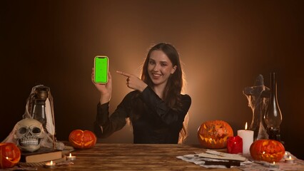 Halloween gothic woman witch fortune teller sitting at table holds smartphone chroma key green screen background pointing finger at it. Magic halloween concept.