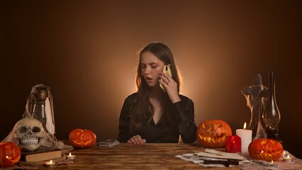 Halloween gothic woman witch fortune teller sitting at table talking on smartphone, annoyed bored face expression. Magic halloween concept.