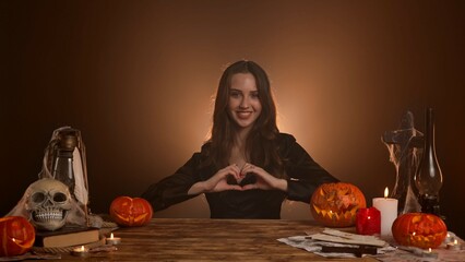 Halloween gothic woman witch fortune teller sitting at table looks around smiles, shows heart hand gesture sign at camera. Magic halloween concept.