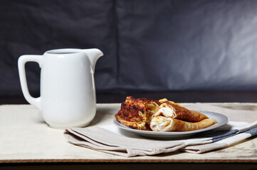 Rolled pancakes with cottage cheese and raisins and Portion of cottage cheese casserole on plate. A delicious sweet dessert and milk jug on kitchen table