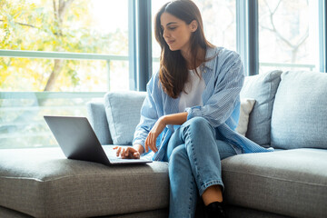 Modern lifestyle of a young woman working remotely from her sofa, dressed in casual denim. She uses wireless technology on her laptop to search for a new job while chatting