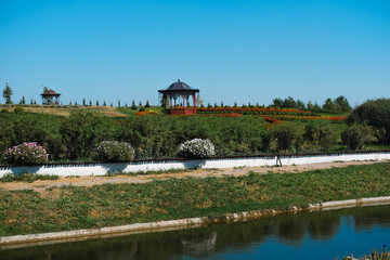 Beautiful garden landscape with gazebos and blooming flowers near a tranquil river on a sunny day