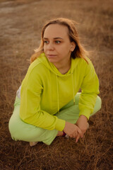 A woman squatting in a bright green hoodie and matching pants on a grassy field during golden hour