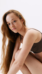 Fototapeta premium Portrait of beautiful young woman with freckled face, long hair, posing in underwear isolated on white studio background. Concept of natural female beauty, body and skin care, health