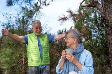 Fototapeta premium Happy carefree senior couple in outdoors excursion in the woods enjoying nature and healthy lifestyle in retirement. Wife holding a coffee cup
