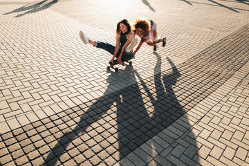 Two happy young women having fun on a skateboard in the city, casting long shadows on a sunny day