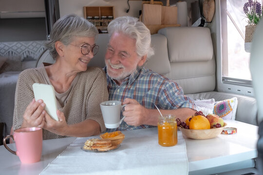 Cheerful senior couple in travel vacation leisure inside a camper van dinette enjoying breakfast together while using smartphone