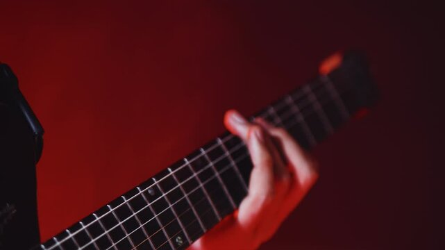Close-up of the hands of the guitarist playing chords on the guitar fretboard while performing a cheerful rock song on the show under red concert lights