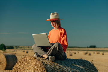 Focused woman wearing hat and using laptop sitting on hay bale in field