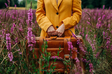 Hands of woman holding suitcase and standing amidst pink flowers in field