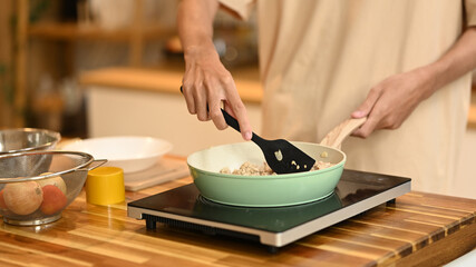 Close up of man using a spatula to stir and cook the ingredients in frying pan on an induction stove