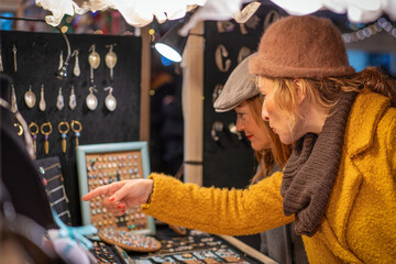 Women shopping jewelry at Christmas market, friends browsing handmade accessories in winter