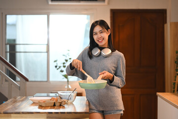 Attractive young woman holding a green frying pan and stirring with a spatula, cooking in a kitchen