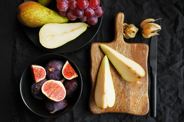 Ripe whole and sliced ​​figs, pears and pink grapes on a wooden board and black plate next to physalis fruits on a dark background
