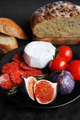Plate with appetizers of Brie cheese, perroni, tomatoes, figs and rosemary sprigs next to sliced ​​grain bread on a dark background