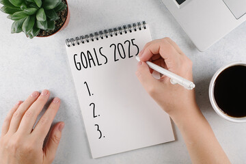 Woman's hand writing 2025 Goals in notebook. Laptop, home plant, cup of coffee on light table. Top view, flat lay