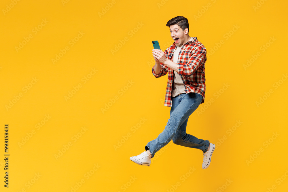 Poster Full body side view young happy man wears red checkered shirt casual clothes jump high hold in hand use mobile cell phone isolated on plain yellow orange background studio portrait. Lifestyle concept.