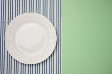 Empty round ceramic plate and blue kitchen towel on green background, top view