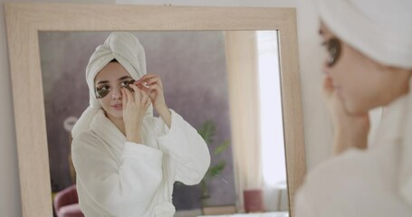 Young beautiful woman in a bathrobe and a towel on head look at the mirror with healthy skin laughs joyfully applies brown patches to reduce eye bags. Cosmetics and anti age plastic surgery.