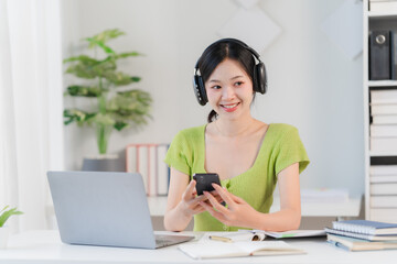 Smiling woman with headphones, working from home on laptop with phone in hand, looking at camera with happy expression 