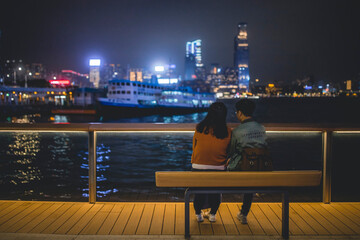 Nighttime scene of a couple sitting on a bench by the waterfront, gazing across the Victoria Harbour in Hong Kong