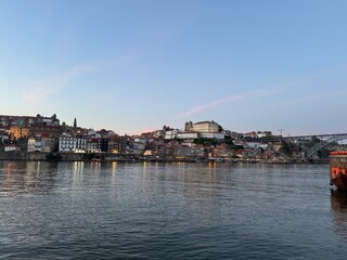 Fototapeta premium An enchanting panorama of Porto's old town as seen from the Gaia side, with traditional Rabelo boats drifting on the Douro River against the backdrop of a breathtaking sunset.