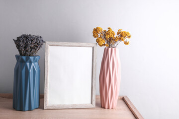 Vases with dry flowers and white blank photo frame on wooden table, gray background