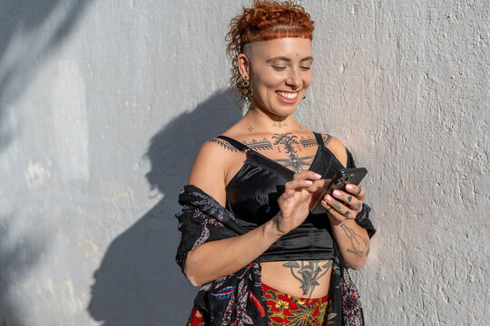 Cheerful woman using cellphone against wall on sunny day