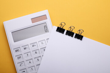 Calculator and white sheet of paper with binders on yellow background. Business concept
