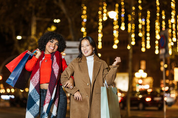 Two young women holding shopping bags and walking down a decorated street at night