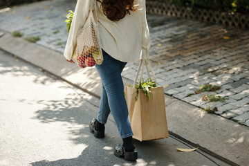 Individual walking down street with groceries in reusable bags, seen holding produce and paper bag....