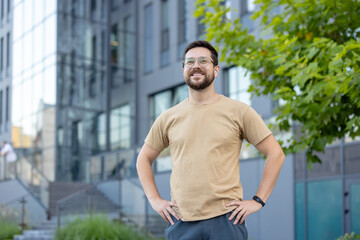 Portrait of a young smiling man standing proudly on a city street in sportswear, holding his hands on his waist and resting contentedly after a run