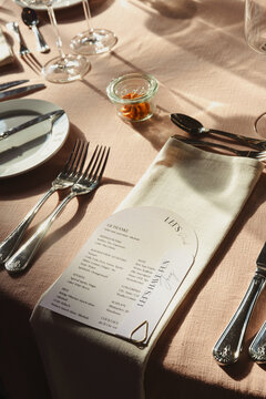 An modern, elegant wedding table setup with a menu, silverware, and glassware arranged on a peach-colored tablecloth under natural sunlight