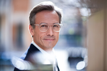 Obraz premium Sophisticated mature man in glasses, wearing a suit, standing confidently outdoors with a blurred urban background