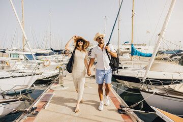 A smiling couple walks hand in hand along the marina's dock, surrounded by boats on a sunny day.