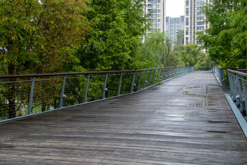 A long wooden pedestrian bridge with metal railings stretches across a public outdoor green space in an urban park, with some modern high-rise residential apartment buildings in the distance. 